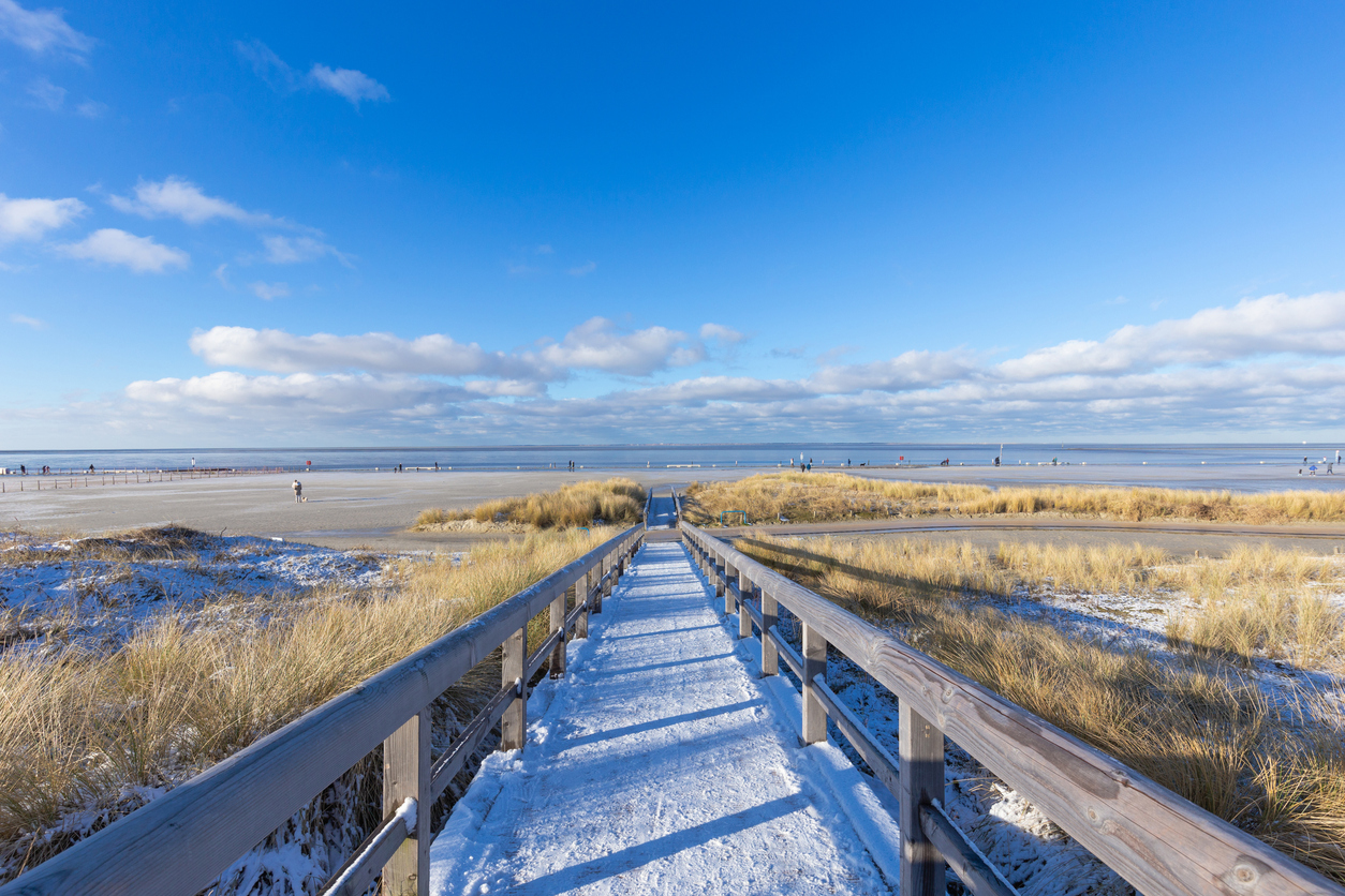 Winterlicher Strandzugang in Norddeich mit verschneitem Holzsteg, Dünen und Blick auf die Nordsee.