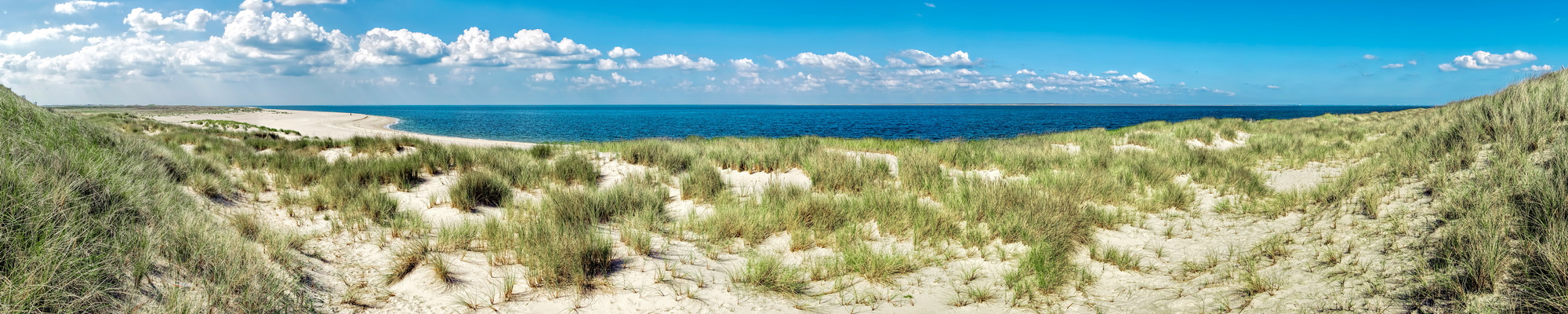 Weites Panorama der Dünenlandschaft auf Norderney mit weißem Sandstrand und tiefblauem Meer unter sommerlichem Himmel. Das Bild zeigt die typische Natur der Insel, die Gäste einer Ferienwohnung auf Norderney täglich vor ihrer Tür erleben können.